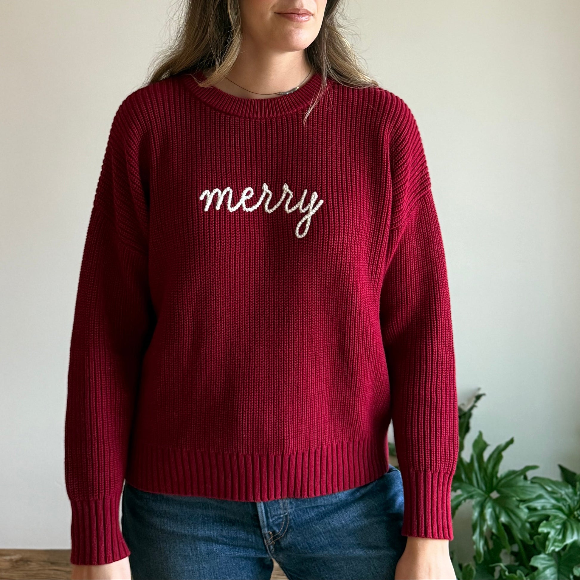 Person wearing a red sweater with 'merry' text against a plain wall.